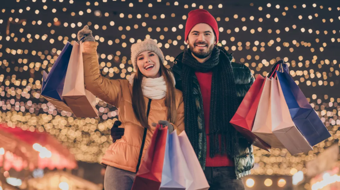 Para w zimowych ubraniach ciesząca się zakupami na tle świątecznych światełek