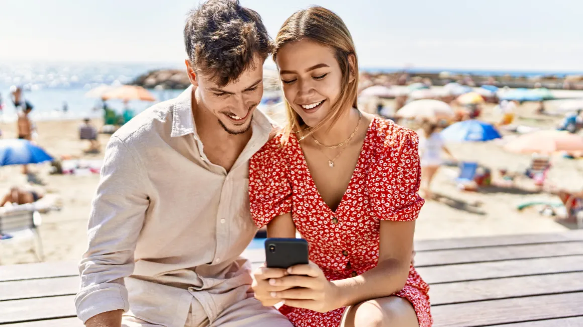 Uśmiechnięci młodzi ludzie nad morzem korzystający z aplikacji na smartfonie
